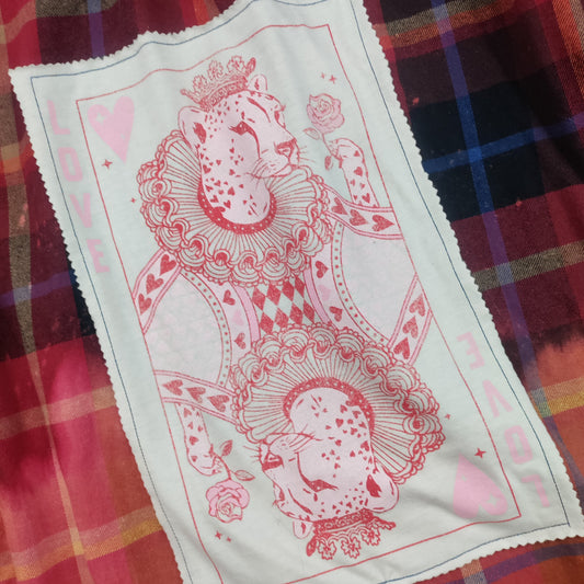 A red and black plaid long-sleeve shirt with a white rectangular patch featuring pink queen of hearts card designs, laid flat on a gray textured blanket.