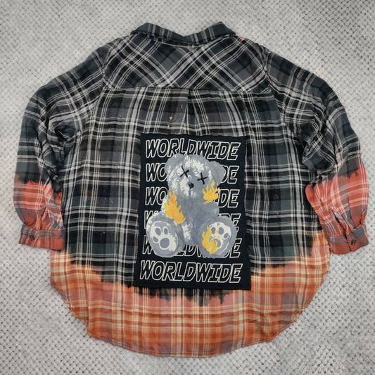 A small plaid shirt laid flat on a gray textured surface. The shirt features a black and white plaid upper section with a central graphic of a fluffy dog and the word 'WORLDWIDE' repeated, and bleached orange and white plaid sleeves and lower hem.