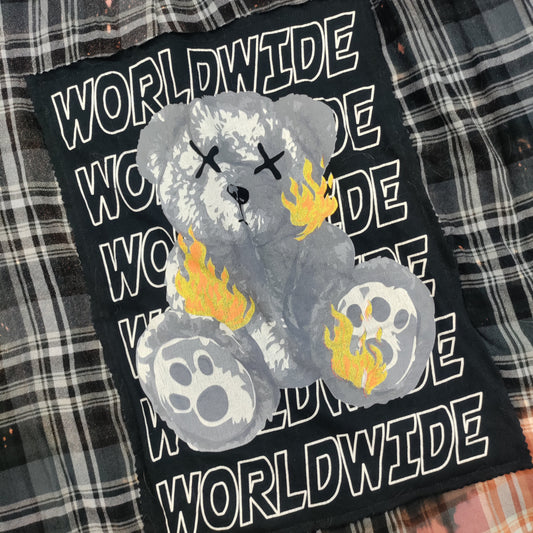 A black patch featuring a distressed gray teddy bear with yellow flames and skull details, surrounded by the word 'WORLDWIDE' in white outlined text, laid flat on a black and white bleached plaid fabric background.