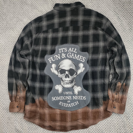A long-sleeve plaid flannel shirt laid flat on a textured gray blanket. The shirt features a black and gray plaid upper body, a brown and tan plaid lower hem, and a large central graphic on the back with a skull and the text 'IT'S ALL FUN & GAMES UNTIL SOMEONE NEEDS AN EYEPATCH'.