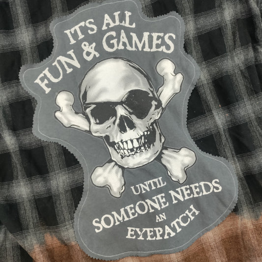 A close-up of a plaid garment featuring a large graphic patch with a skull wearing a black eyepatch and crossed bones, with the text 'ITS ALL FUN & GAMES UNTIL SOMEONE NEEDS AN EYEPATCH'.