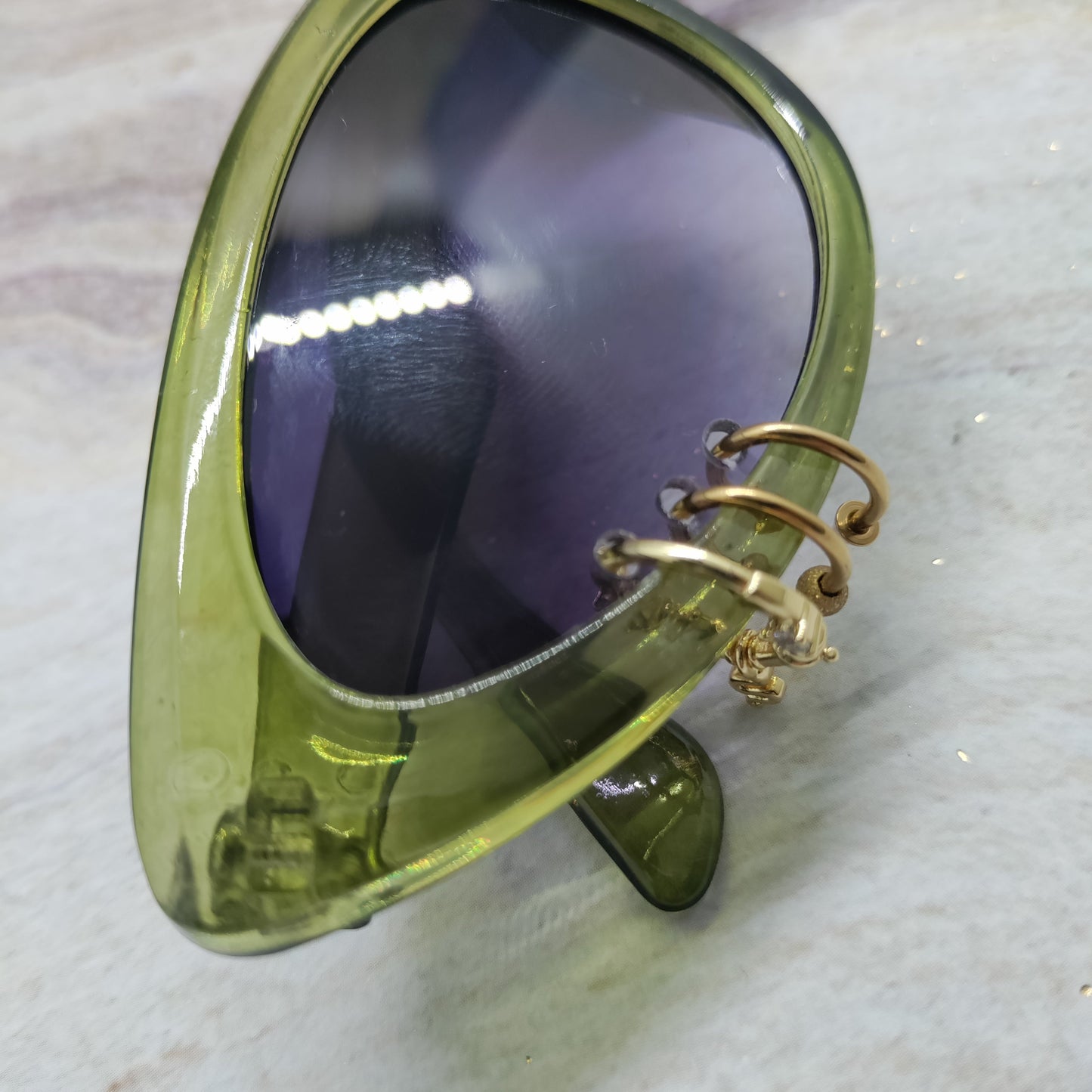 Close-up of a translucent olive green cat-eye sunglasses frame with dark tinted lenses and a gold-tone spiral ear cuff attached on the outer side, resting on a light marble-like surface.