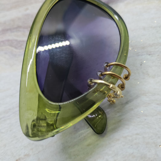 Close-up of a translucent olive green cat-eye sunglasses frame with dark tinted lenses and a gold-tone spiral ear cuff attached on the outer side, resting on a light marble-like surface.