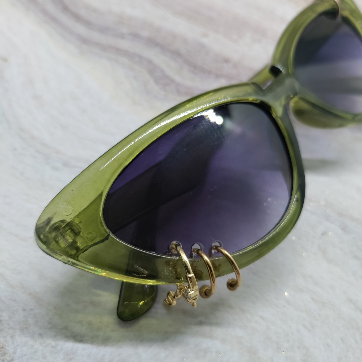 Oversized cat-eye sunglasses with a translucent olive green frame and dark purple tinted lenses, featuring decorative gold wire hoop earrings hanging from the lower frame, resting on a light marble surface.