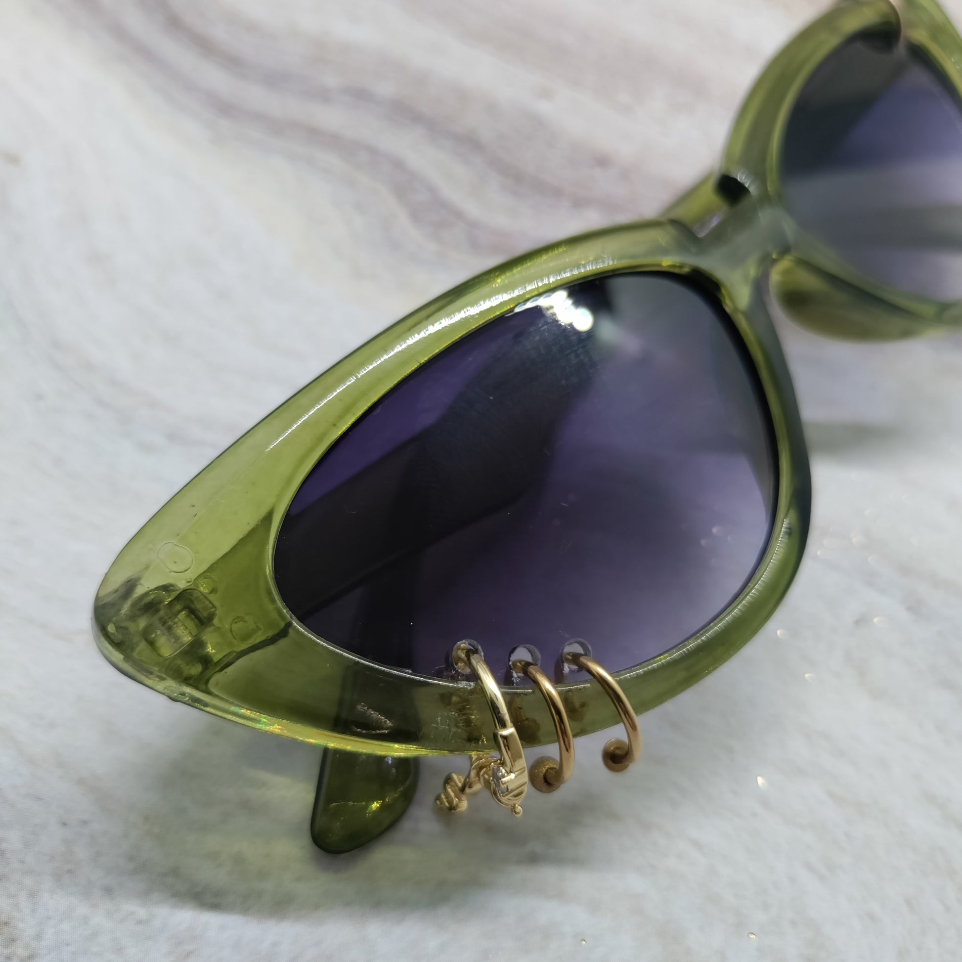 Oversized cat-eye sunglasses with a translucent olive green frame and dark purple tinted lenses, featuring decorative gold wire hoop earrings hanging from the lower frame, resting on a light marble surface.
