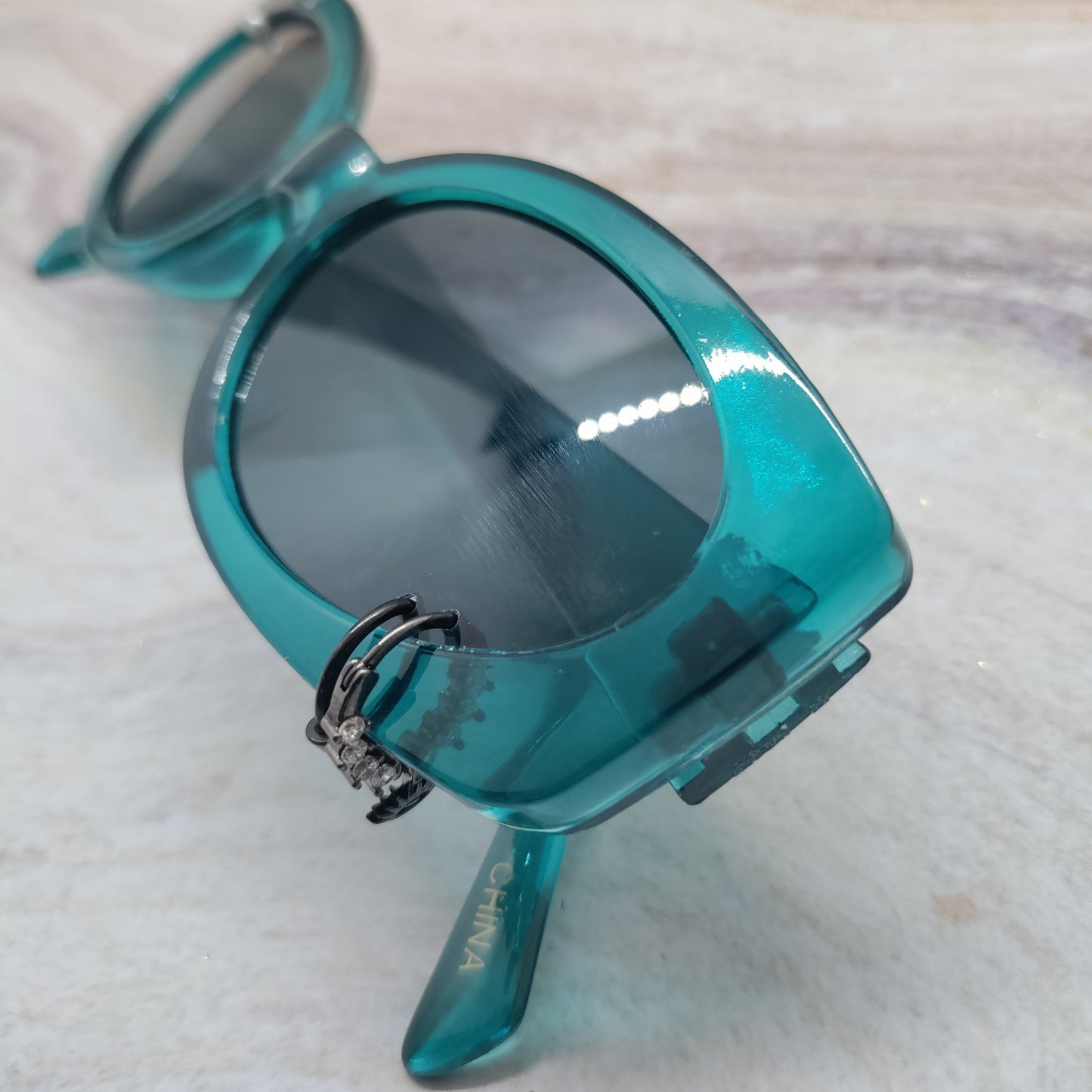 A pair of oversized sunglasses with a translucent teal-blue frame and dark tinted lenses, resting on a light marble surface.