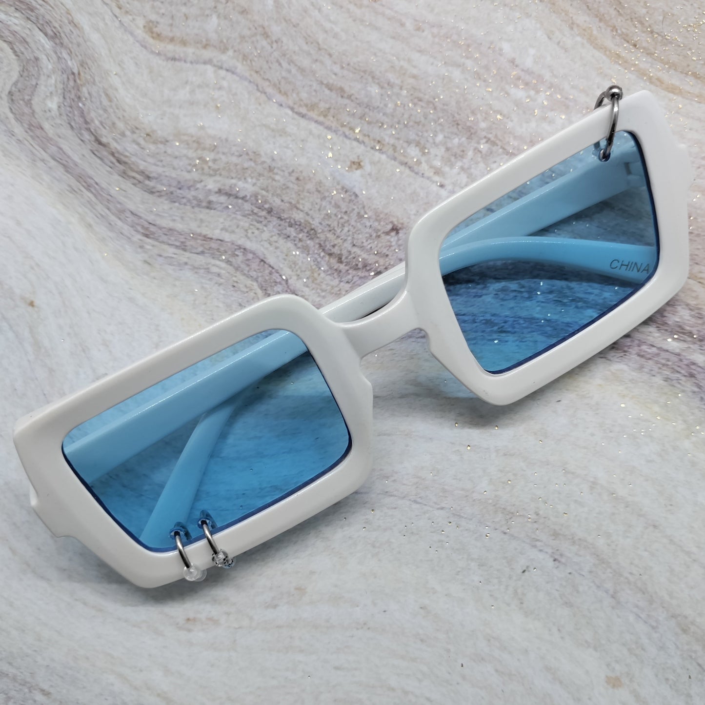 White square sunglasses with light blue lenses resting on a light gray and white marbled surface.