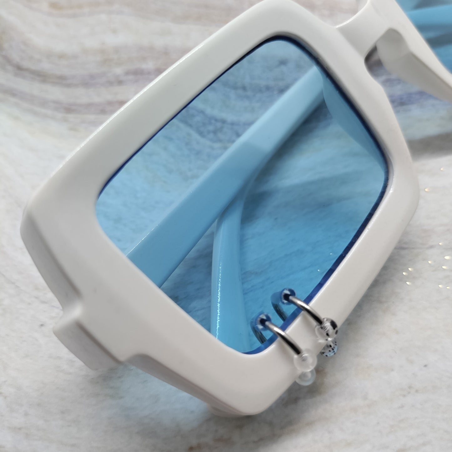 White square sunglasses with light blue tinted lenses and silver metal hinge details, resting on a light marble surface.
