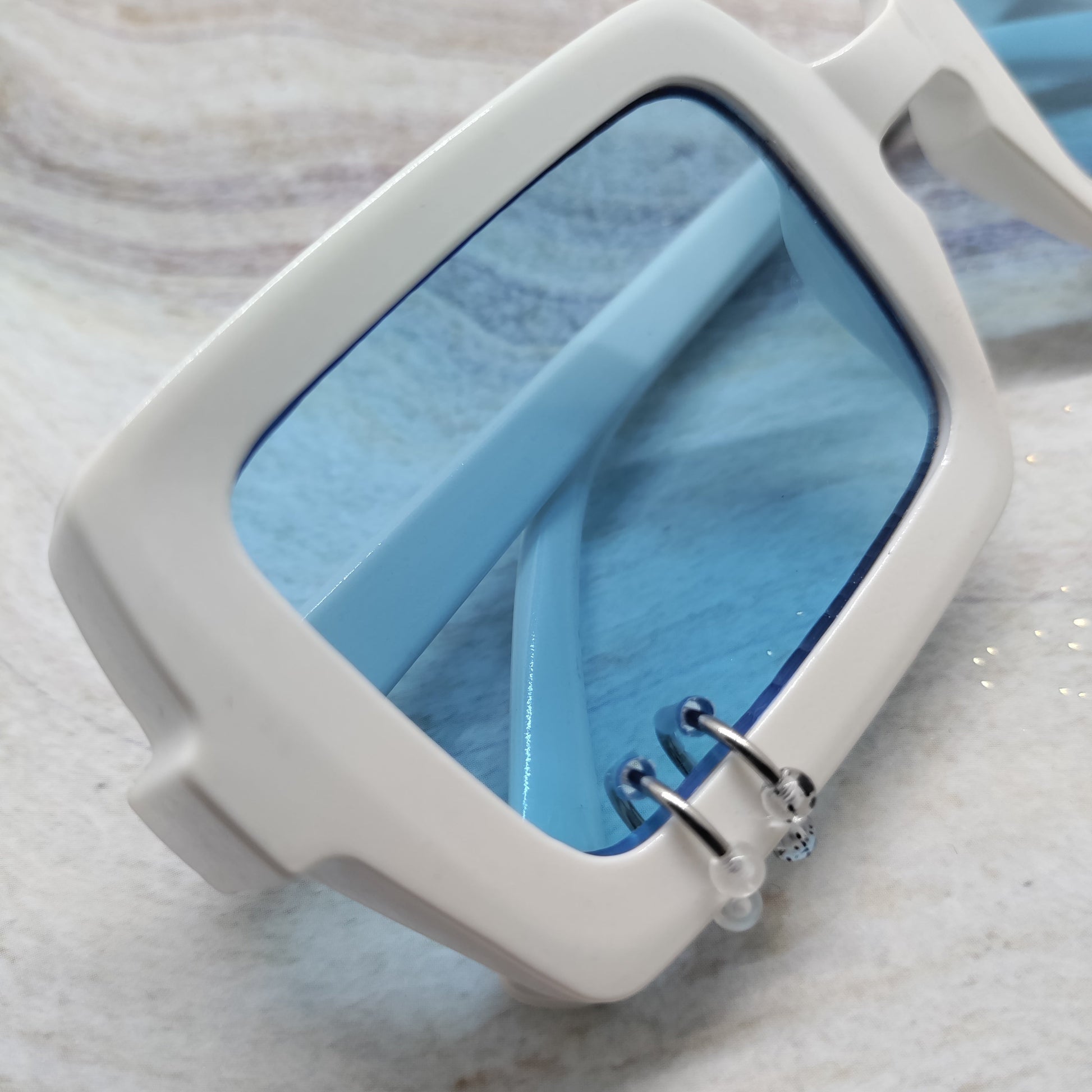 White square sunglasses with light blue tinted lenses and silver metal hinge details, resting on a light marble surface.