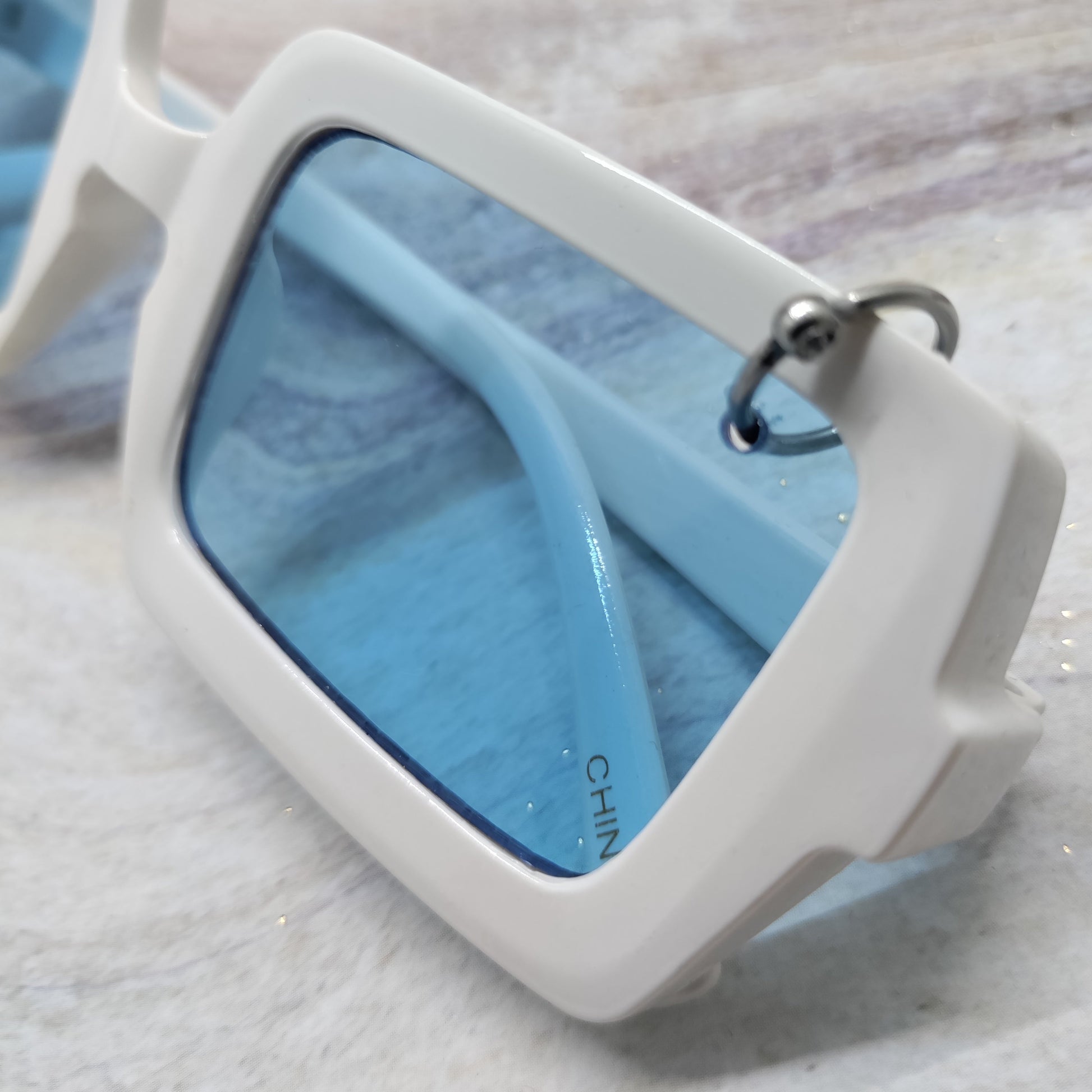 White square-frame sunglasses with light blue lenses, shown close-up on a light textured surface.