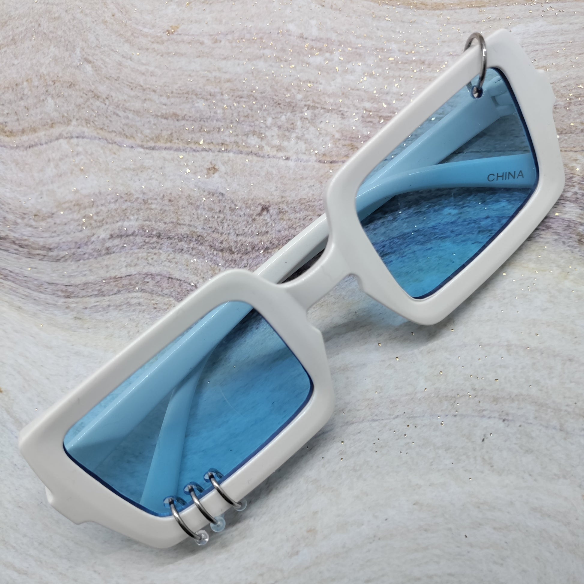 White square sunglasses with blue lenses and small silver metal rings on the front, displayed on a light marble-textured surface.