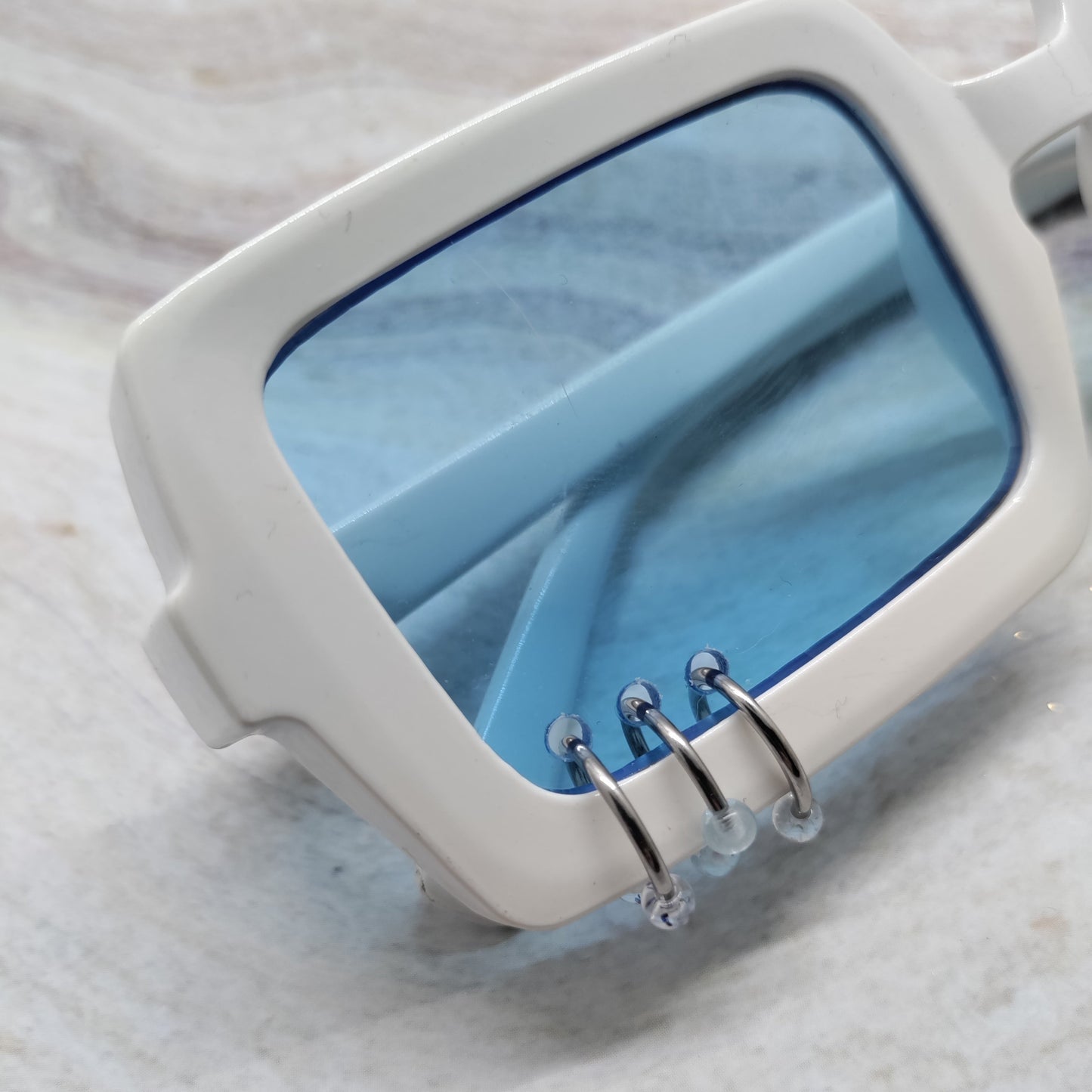 White square sunglasses with translucent light blue lenses and silver metal wire details on the front, resting on a light marble surface.
