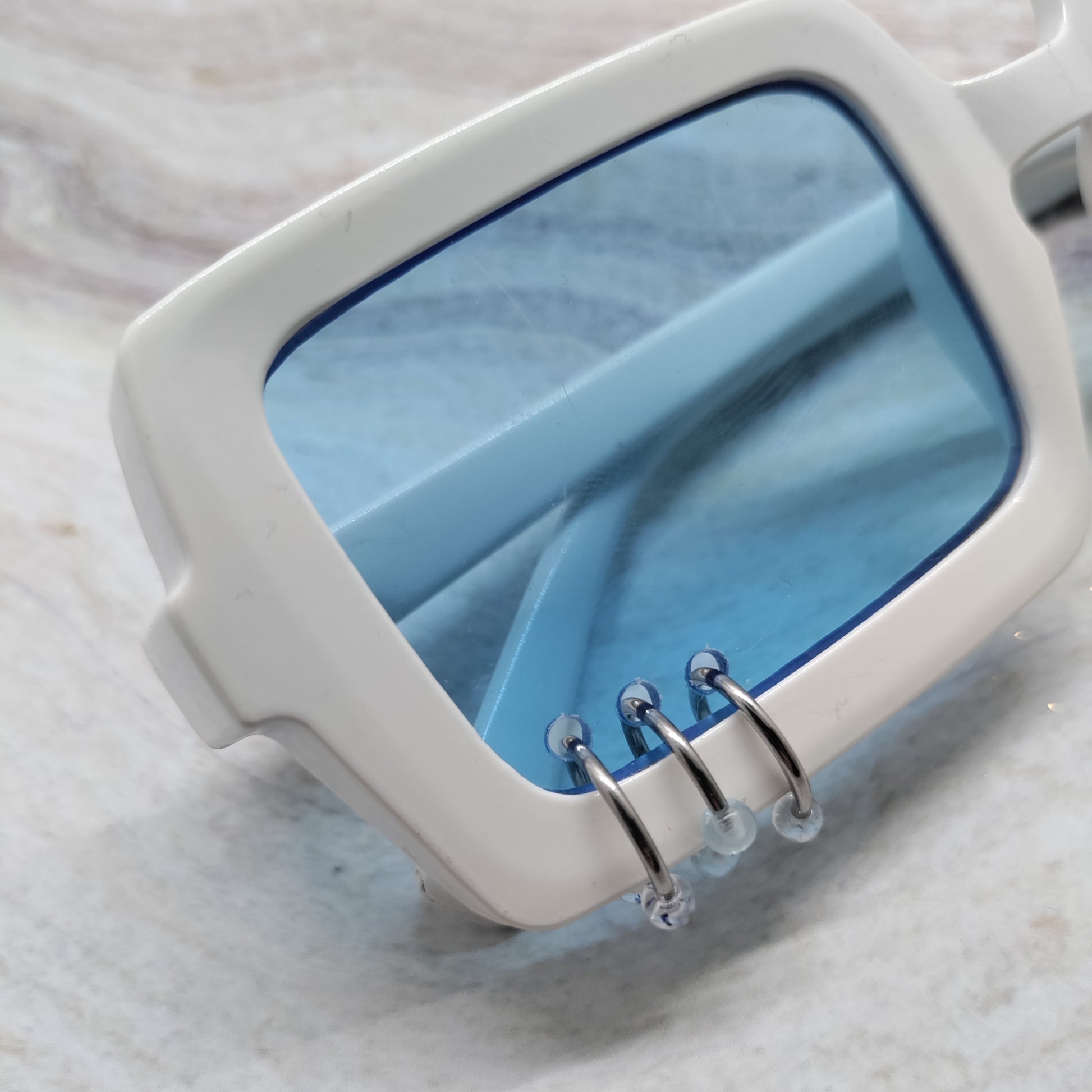 White square sunglasses with translucent light blue lenses and silver metal wire details on the front, resting on a light marble surface.