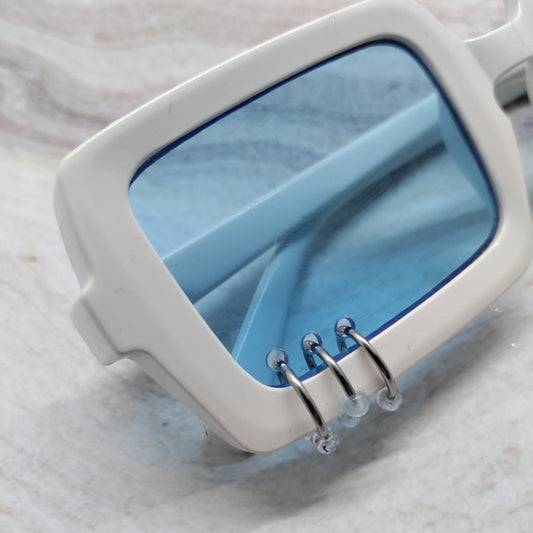 White square sunglasses with translucent light blue lenses and silver metal wire details on the front, resting on a light marble surface.