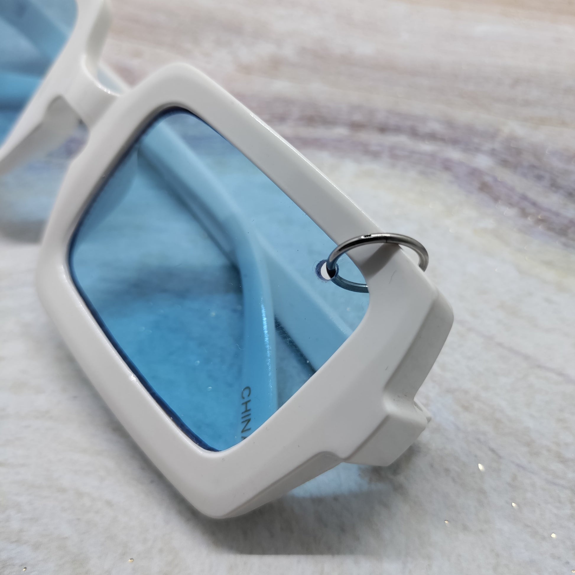 Close-up of a pair of sunglasses with a thick white frame and light blue lenses, resting on a textured light gray surface.