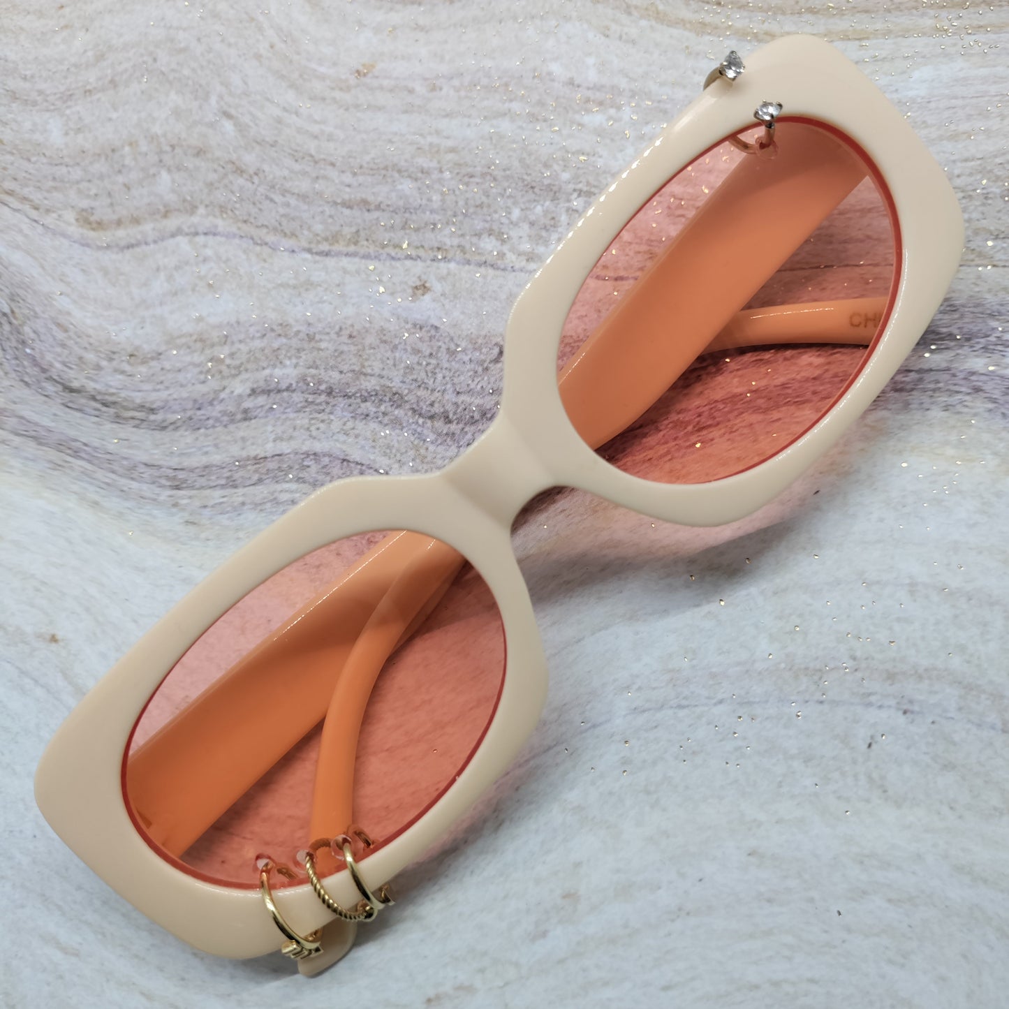 A pair of oversized square sunglasses with a thick cream-colored frame and warm peach-orange lenses, resting on a light gray and white marbled surface.