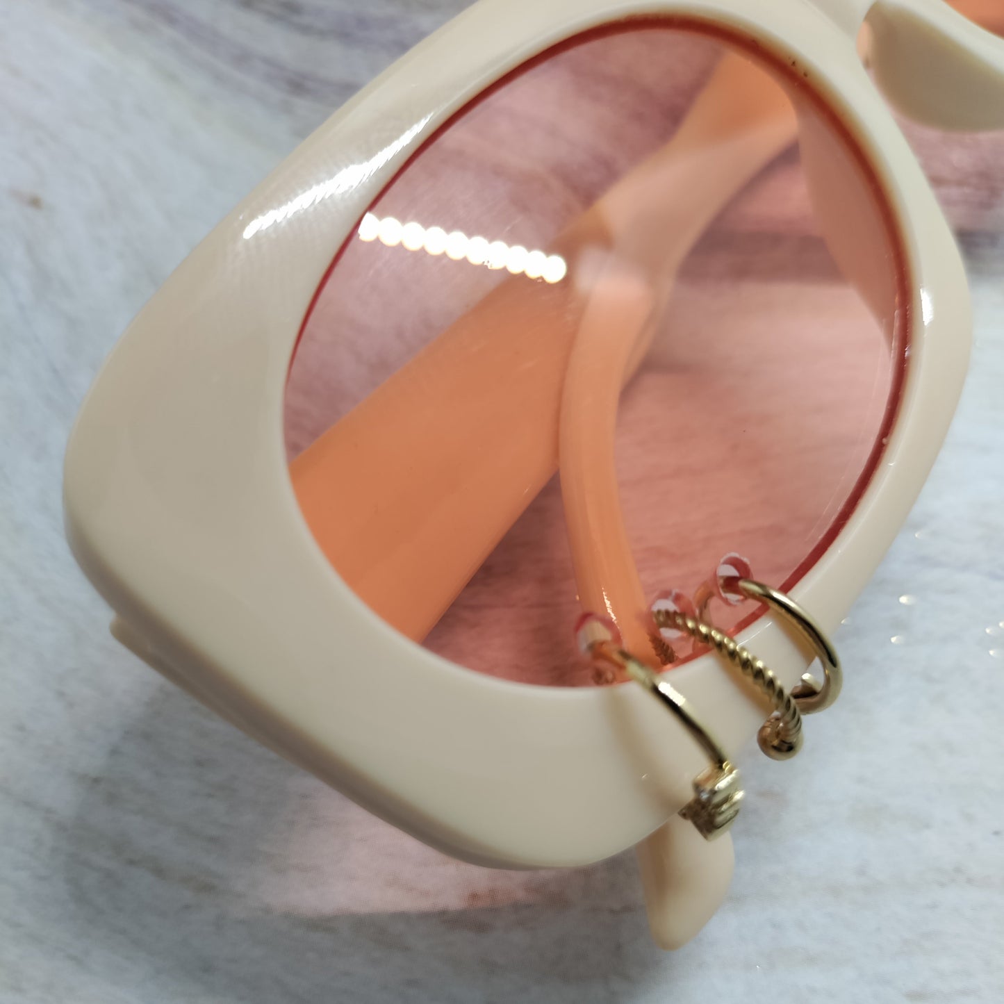 Close-up of a pair of oversized square sunglasses with a thick cream-colored frame, peach-tinted lenses, and gold hoop earrings hanging from the temple.
