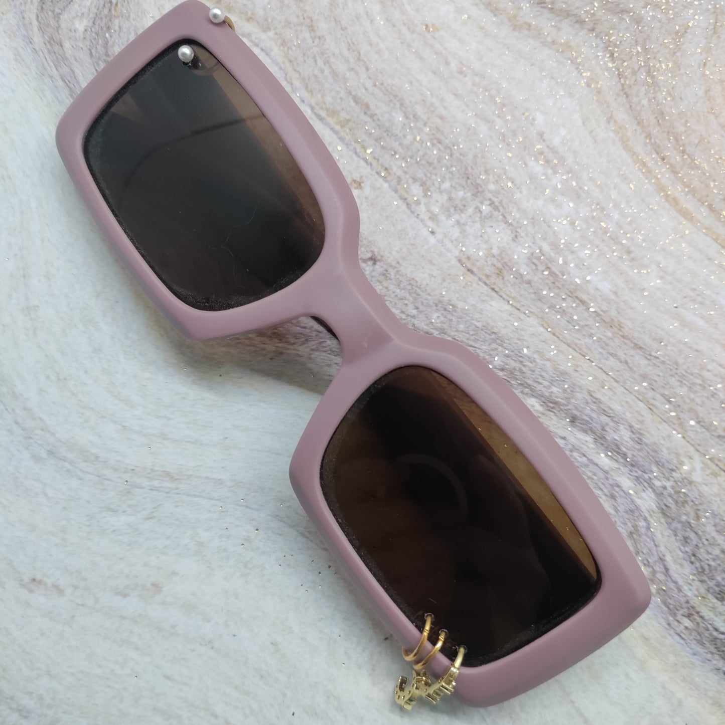 A pair of rectangular sunglasses with a dusty pink frame and dark lenses, featuring a small gold deer head charm hanging from the lower right corner, displayed on a glittery marble surface.