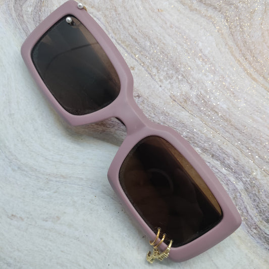 A pair of rectangular sunglasses with a dusty pink frame and dark lenses, featuring a small gold deer head charm hanging from the lower right corner, displayed on a glittery marble surface.