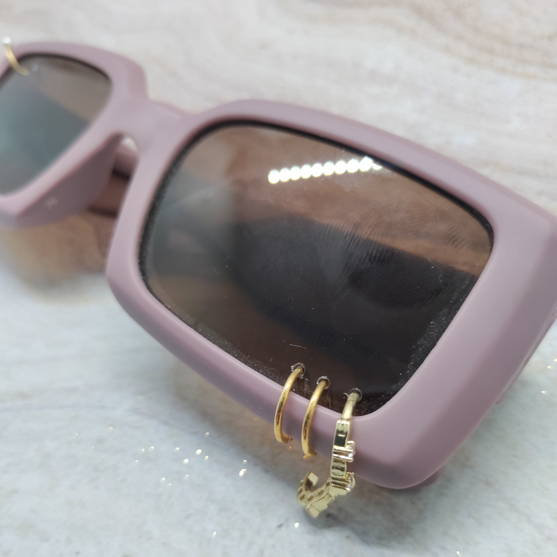 A close-up of dusty pink square sunglasses with dark tinted lenses and gold hoop earrings hanging from the temple, resting on a light marble surface.
