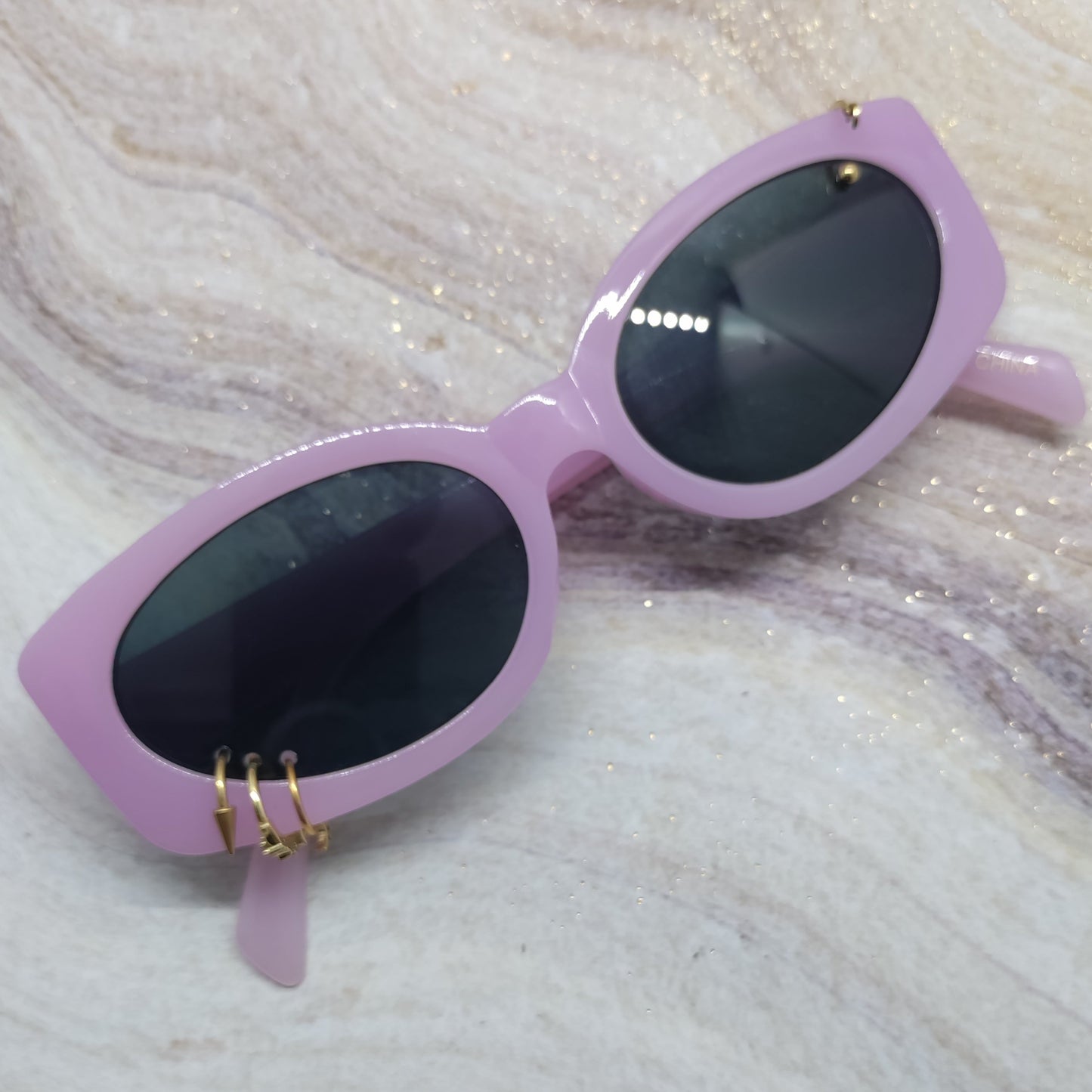 Light purple cat-eye sunglasses with dark lenses and small gold metal accents on the hinge, resting on a white and gray marbled surface with subtle glitter.