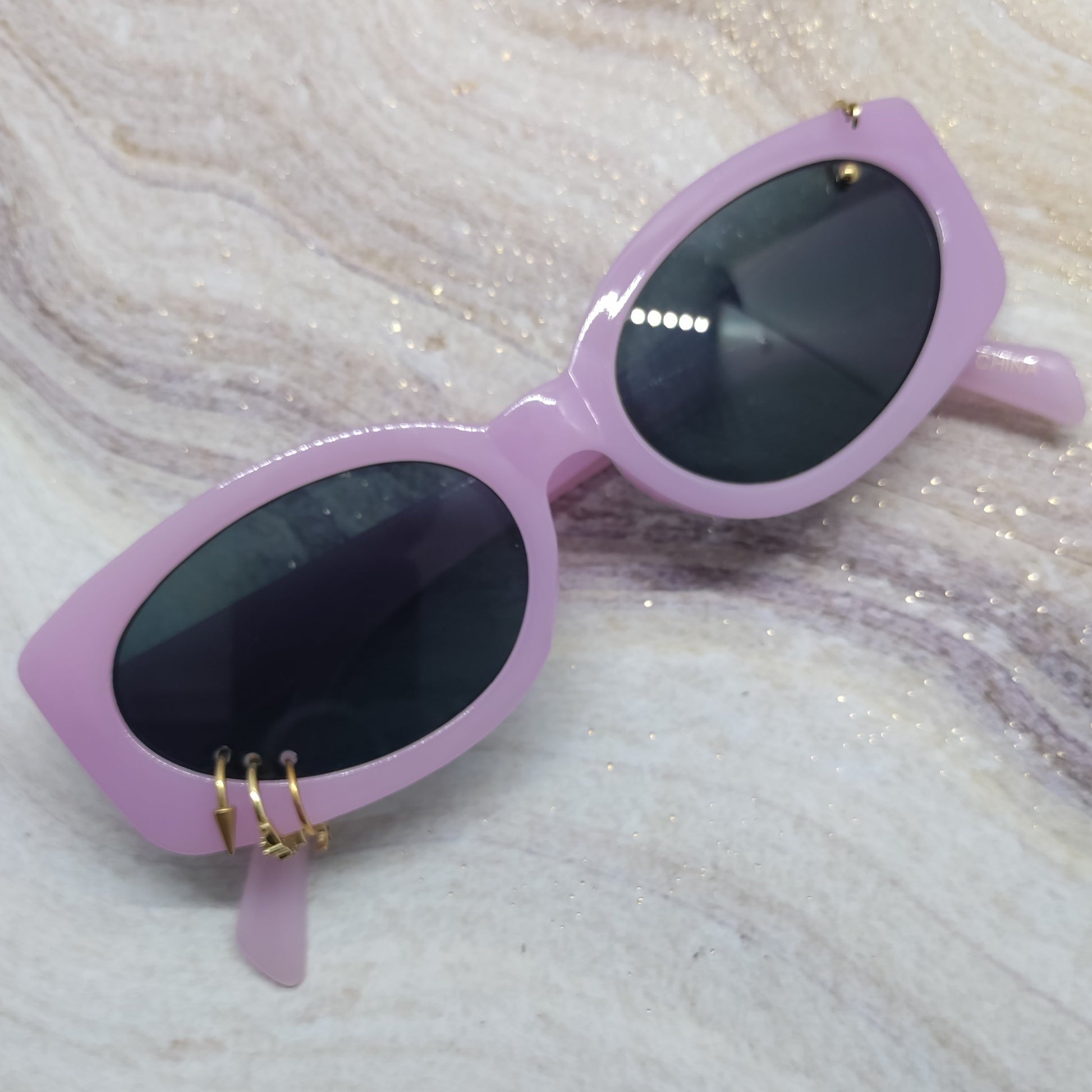 Light purple cat-eye sunglasses with dark lenses and small gold metal accents on the hinge, resting on a white and gray marbled surface with subtle glitter.