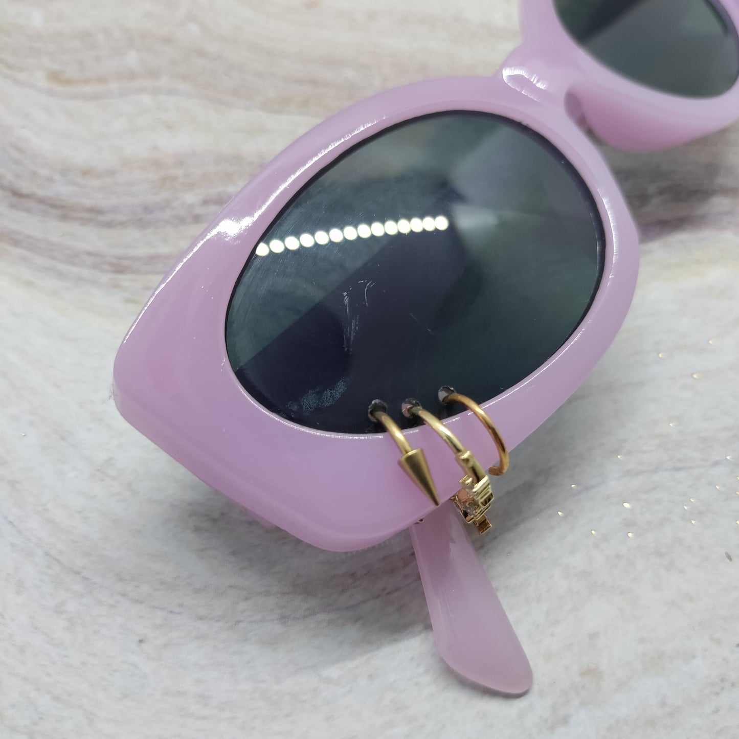 A pair of retro-style sunglasses with a glossy light purple frame and dark lenses, resting on a light marble surface. The left temple arm features a decorative gold-tone ring detail.