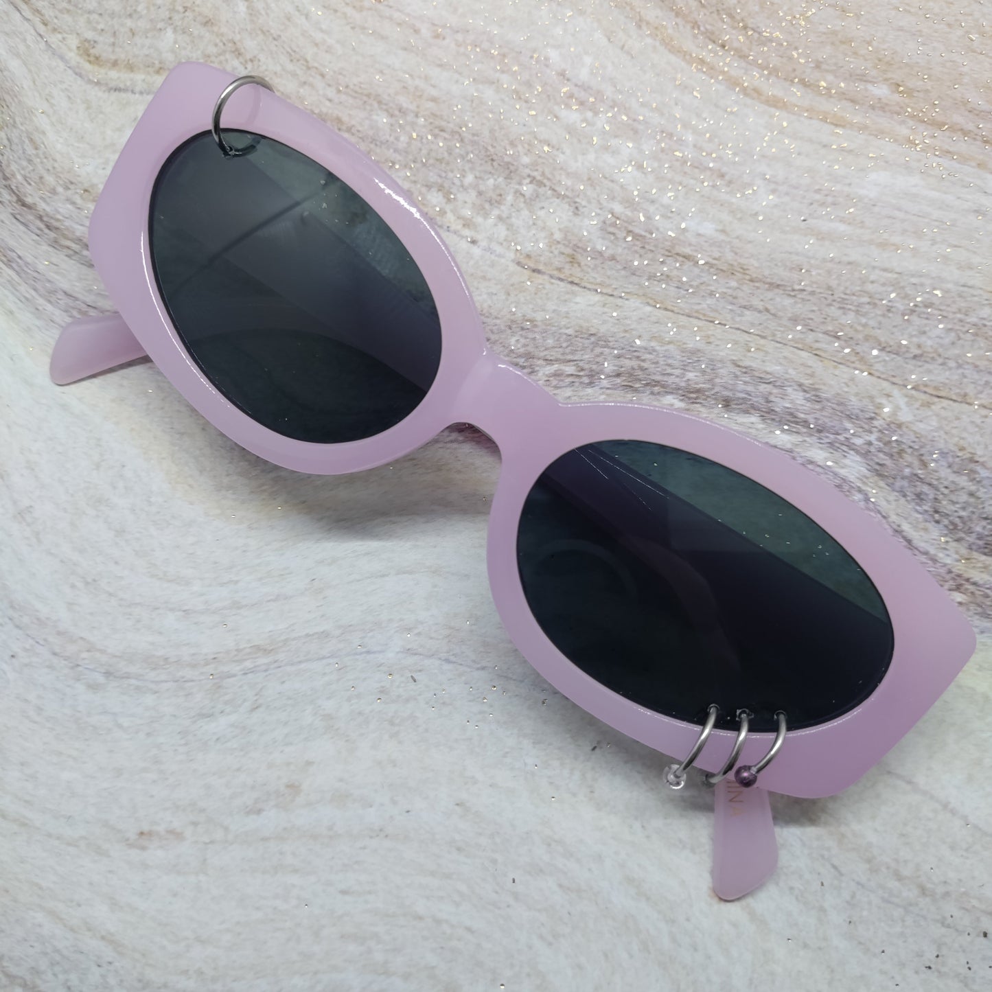 Light purple cat-eye sunglasses with dark lenses and silver hoop earrings, resting on a textured beige marble surface.