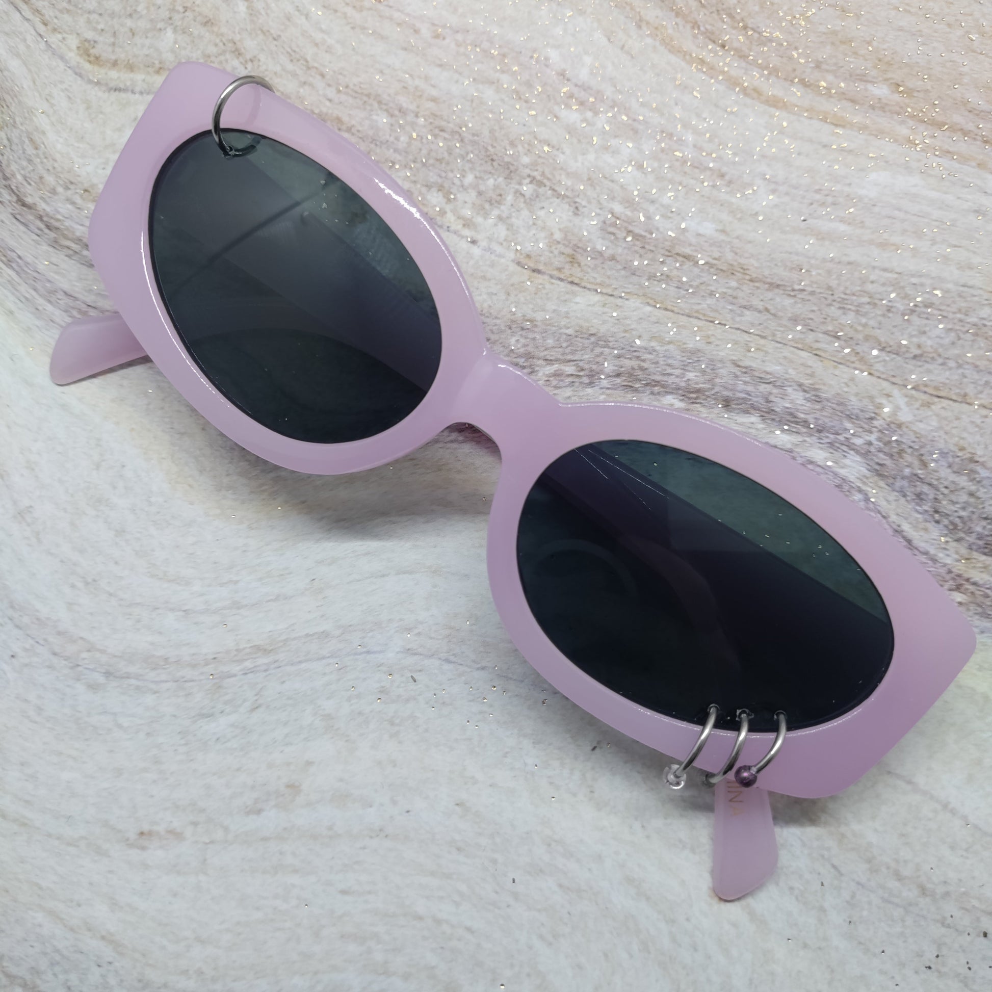 Light purple cat-eye sunglasses with dark lenses and silver hoop earrings, resting on a textured beige marble surface.