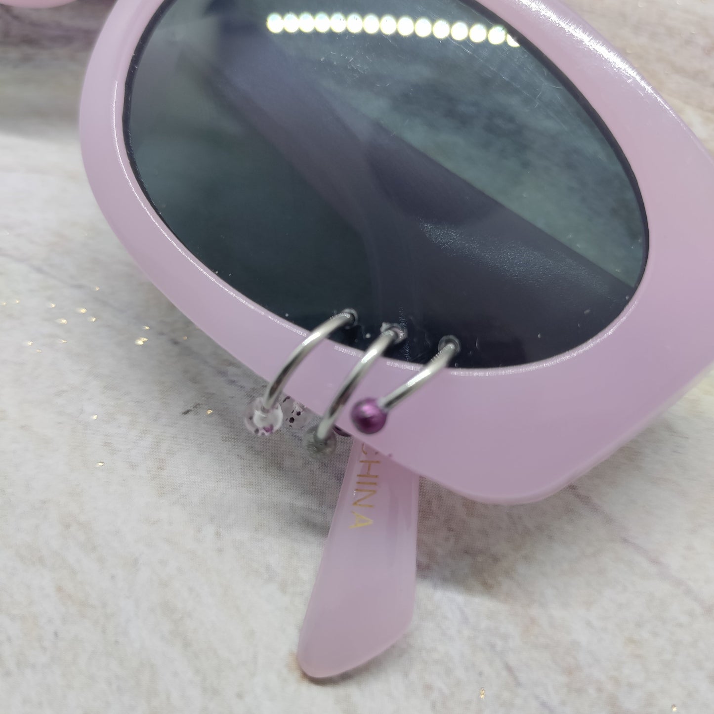 Pink cat-eye sunglasses with dark tinted lenses and a small purple rhinestone accent on the hinge, resting on a light marble surface.