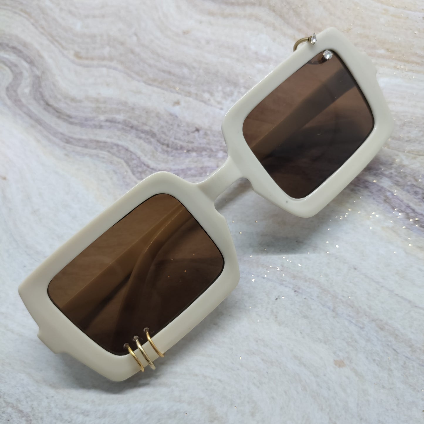 Rectangular sunglasses with thick off-white frames and dark brown tinted lenses, featuring a small gold accent on the hinge, displayed on a light marble surface.