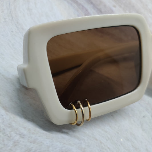 A pair of oversized square sunglasses with a thick cream-colored frame, dark brown lenses, and three small gold rings hanging from the bottom edge, resting on a white textured surface.