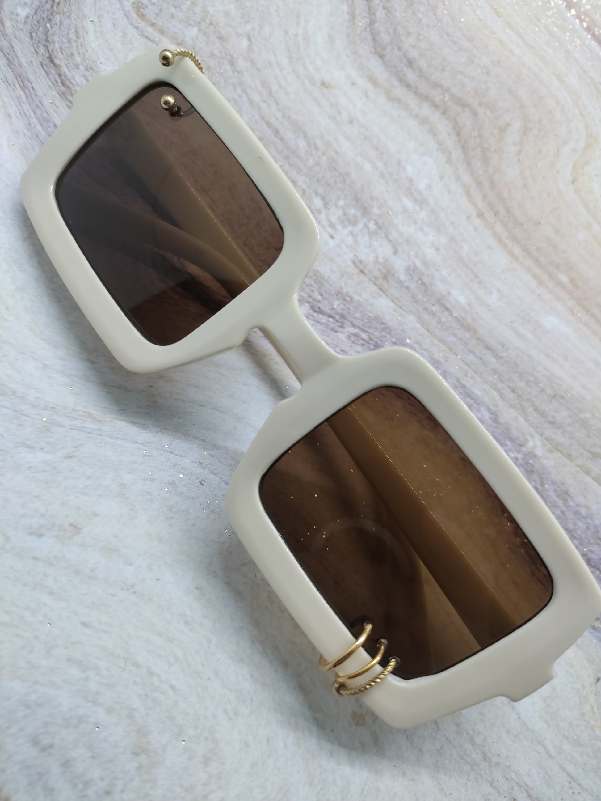 Square-frame sunglasses with thick off-white frames and dark brown lenses, featuring gold metal accents on the hinges, displayed on a light marble surface.