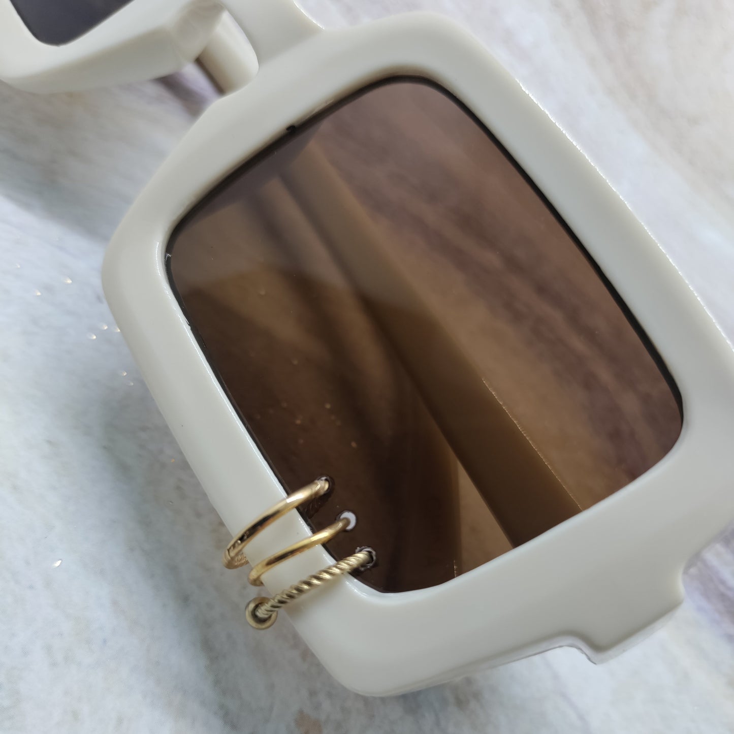 Close-up of a pair of sunglasses with a thick off-white frame and a brown tinted lens, featuring a decorative gold wire ring detail on the side.