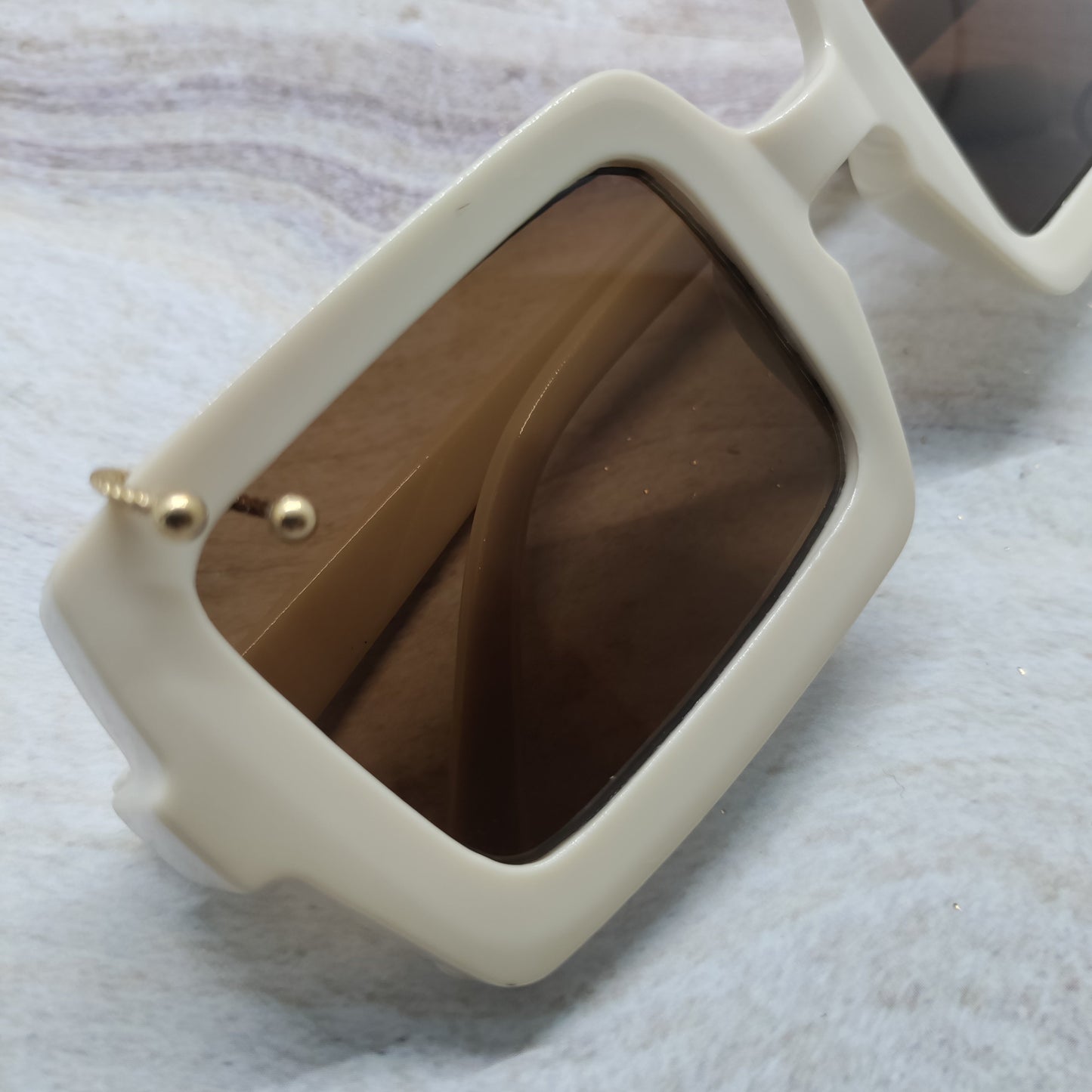A close-up of a pair of oversized square sunglasses with a thick cream-colored frame and dark brown tinted lenses, resting on a light gray marbled surface.