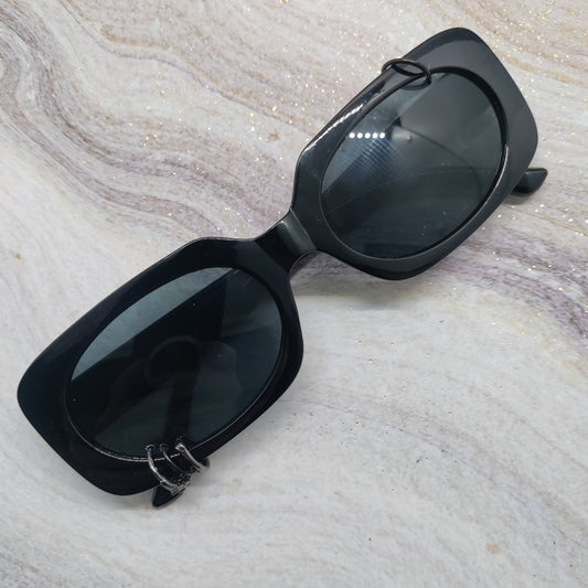 Black square sunglasses with dark tinted lenses and a small silver ring detail on the lower left frame, displayed on a light beige marble surface.