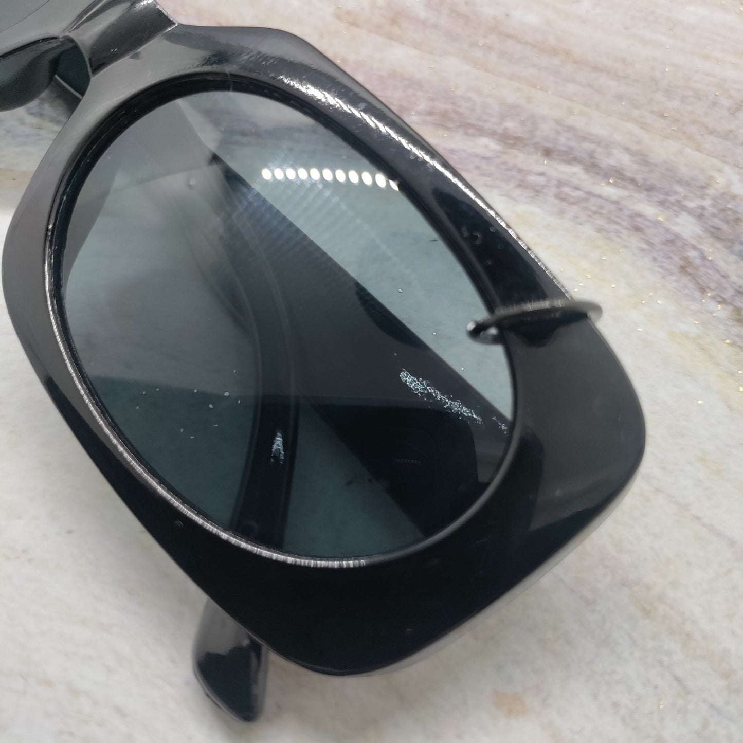 A close-up of a black sunglasses frame with dark tinted lenses, resting on a light beige marble surface.