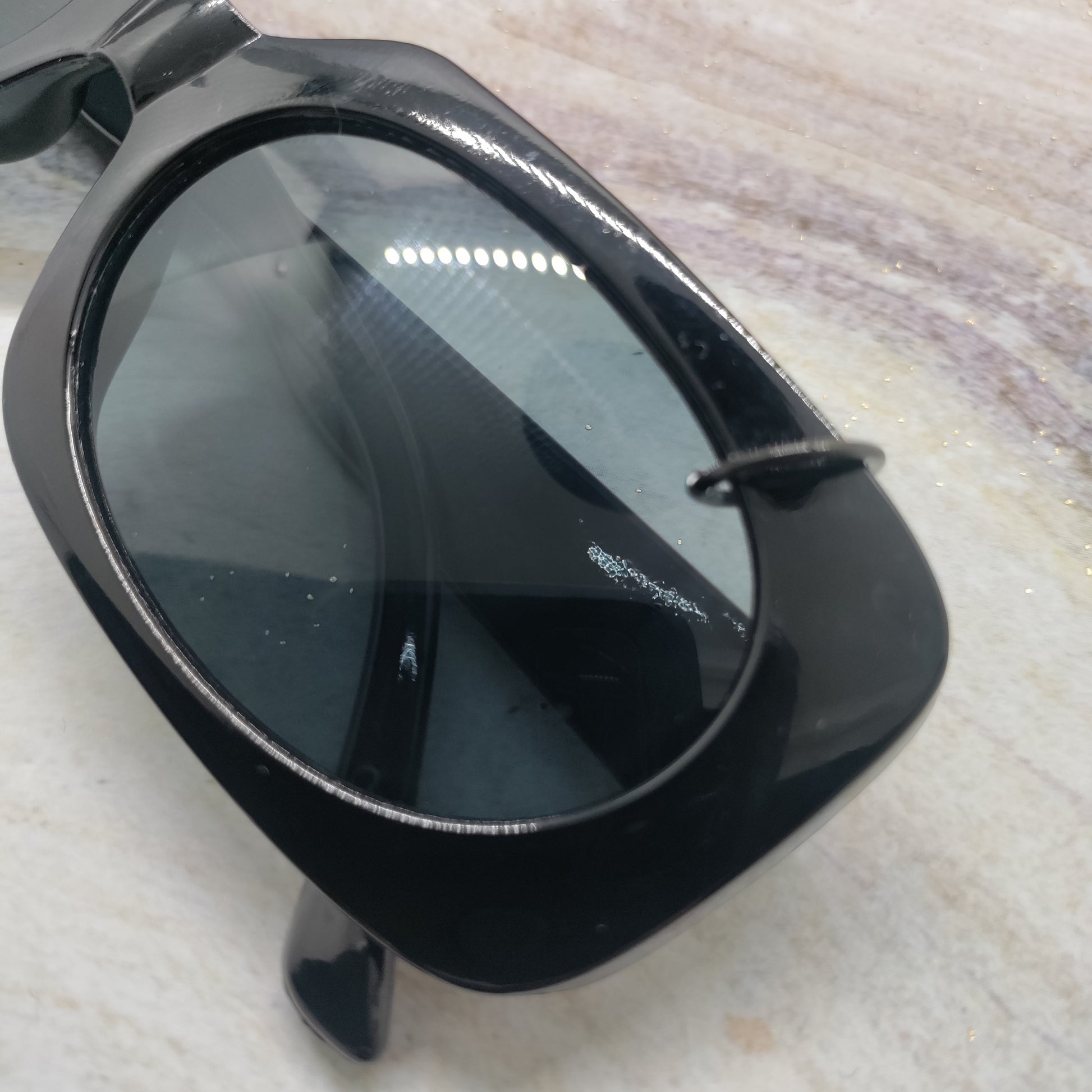 A close-up of a black sunglasses frame with dark tinted lenses, resting on a light beige marble surface.