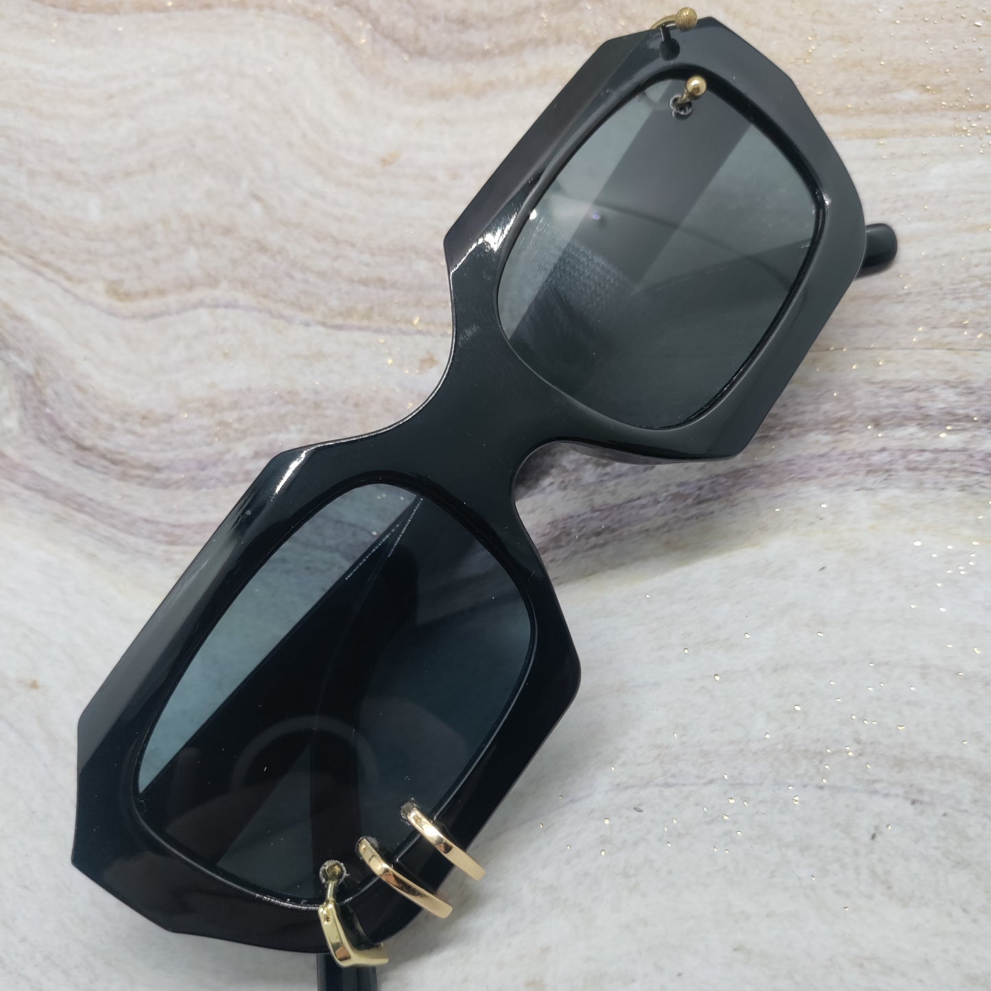 Black oversized square sunglasses with dark tinted lenses and gold hoop earrings hanging from the temples, displayed on a light marble-patterned surface.