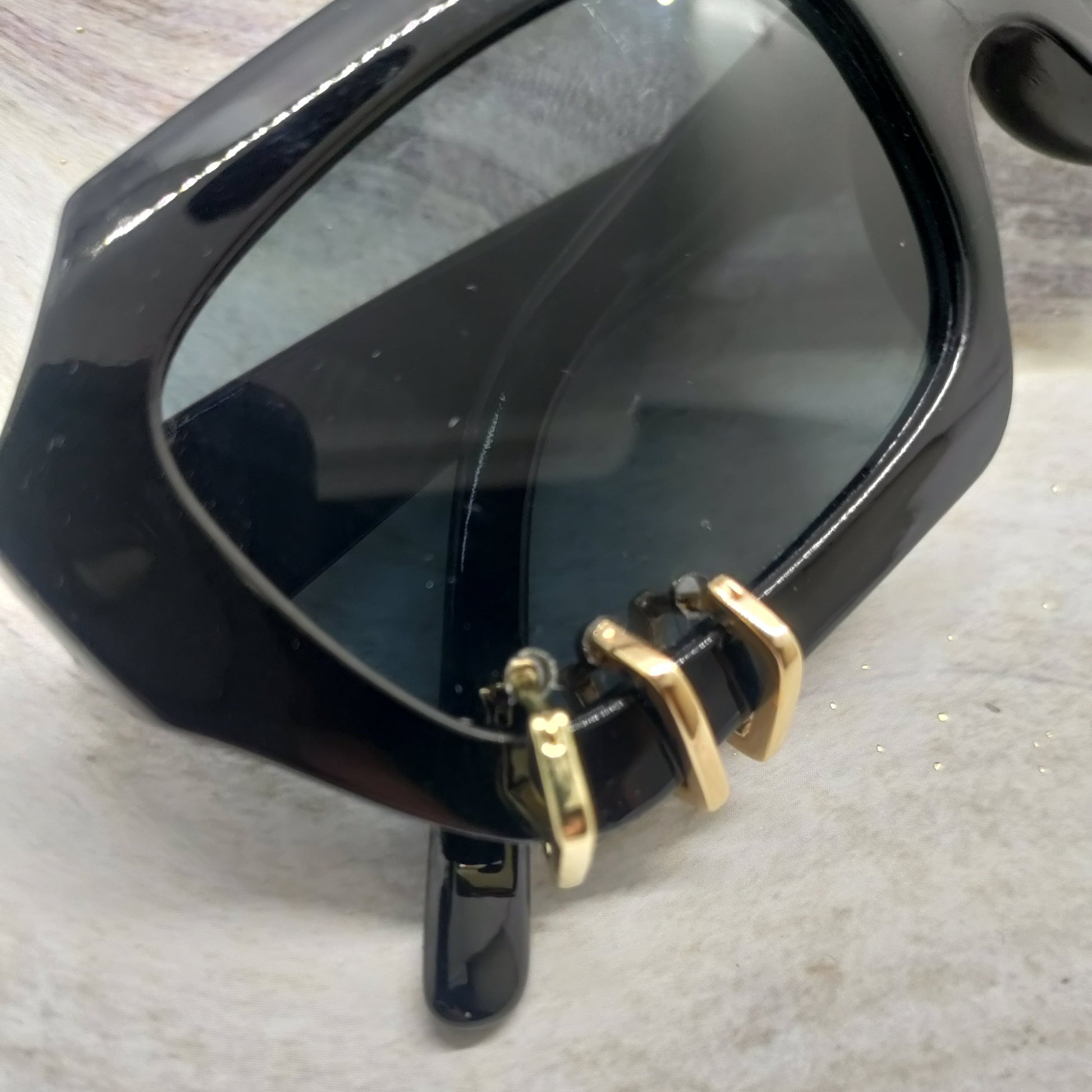Close-up of a black acetate sunglasses frame with dark tinted lenses and gold metal hinge hardware, resting on a light textured surface.