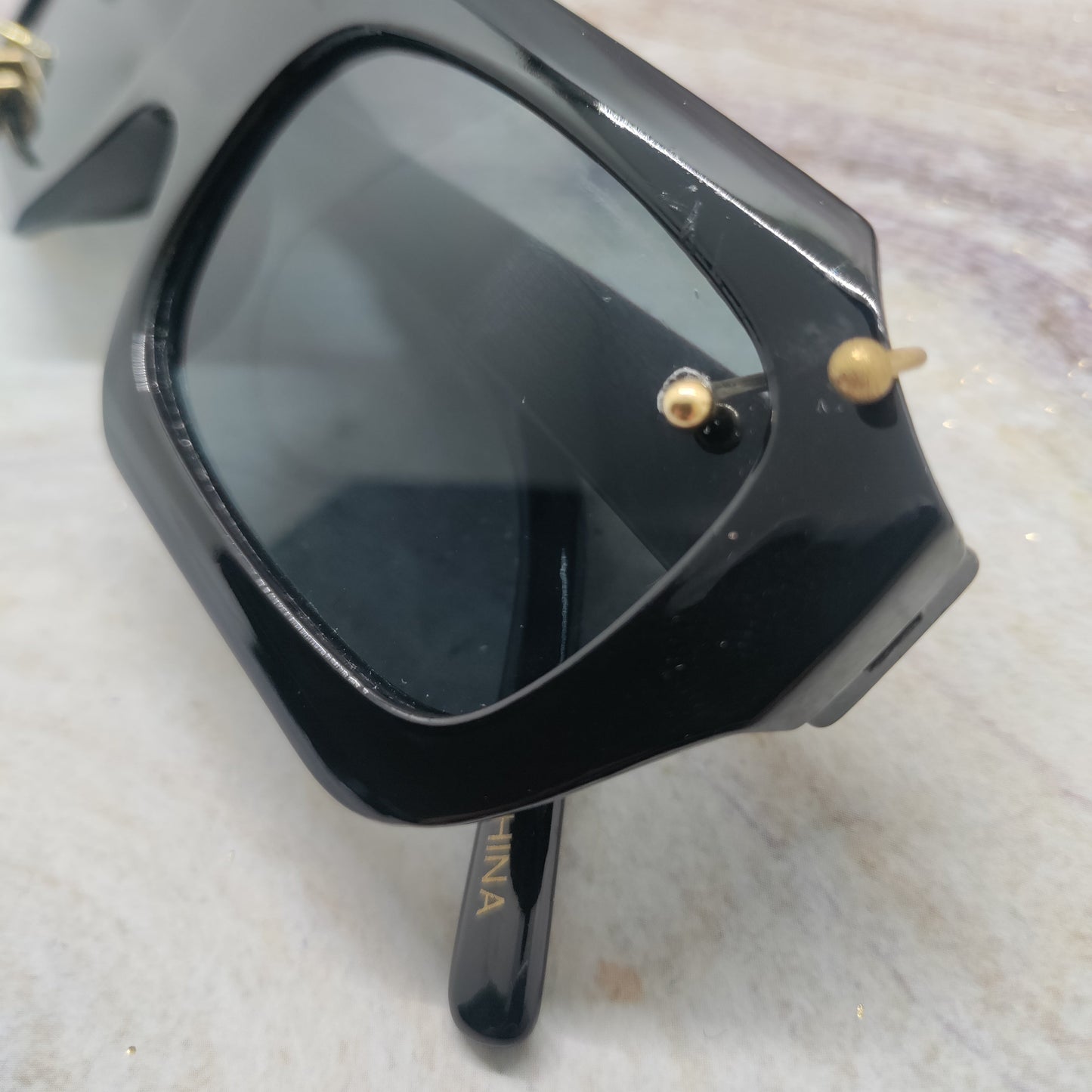 Black square sunglasses with dark lenses and small gold-tone decorative accents on the frame, photographed on a light marble-like surface.