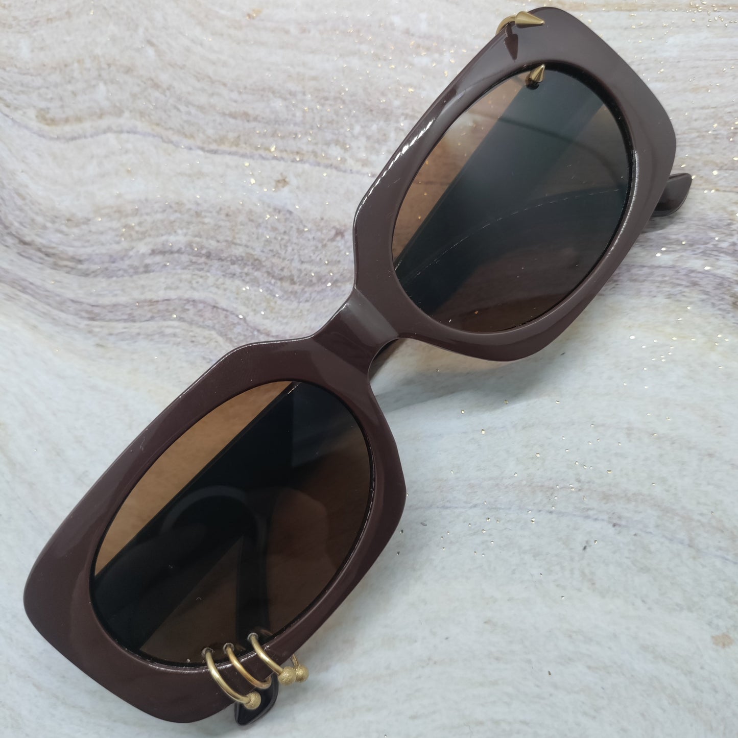 Brown cat-eye sunglasses with dark lenses and small gold metal accents, resting on a light marble-patterned surface.