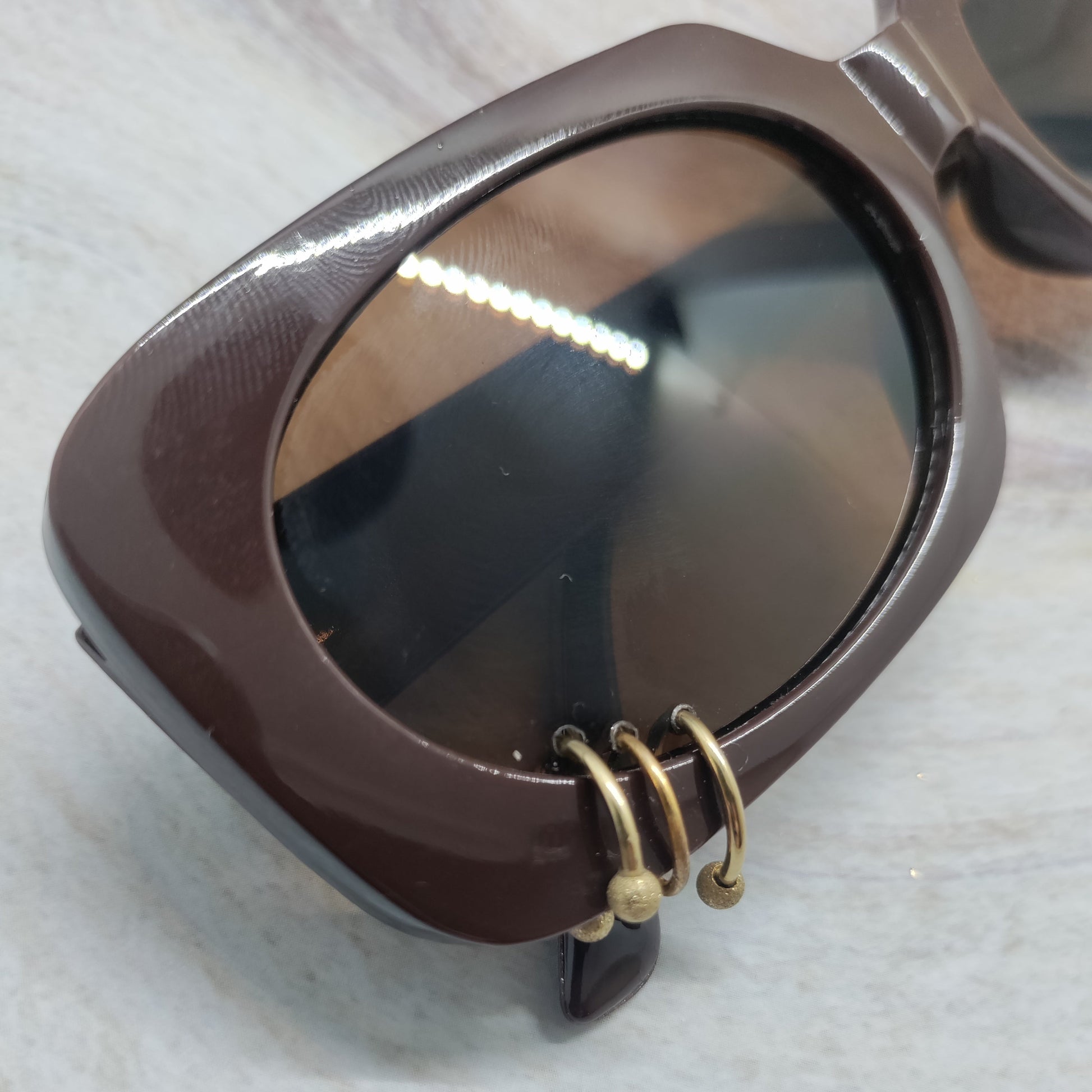 A close-up of a brown acetate sunglasses frame with a unique gold-tone spiral ring detail on the lower edge of the lens, resting on a light marble surface.
