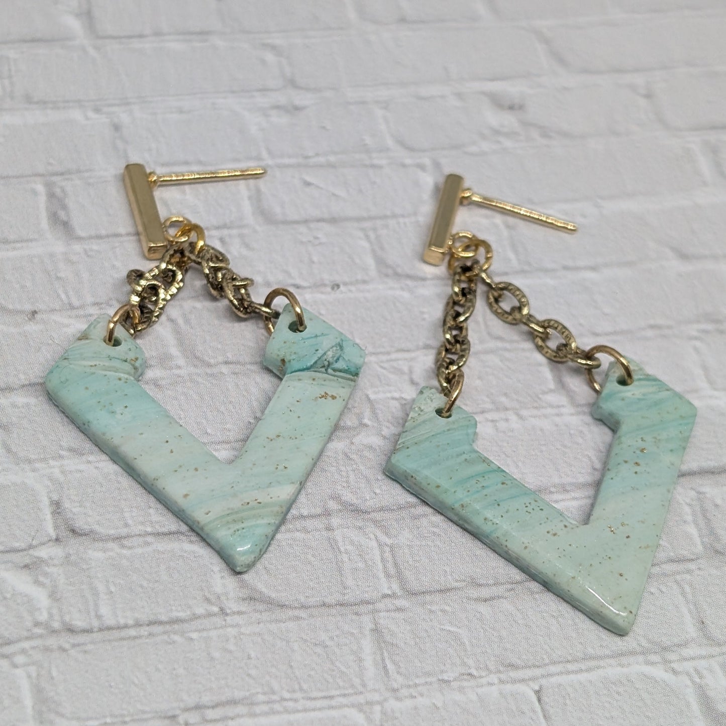 Pair of earrings with gold chains and light blue geometric shapes on a white brick background