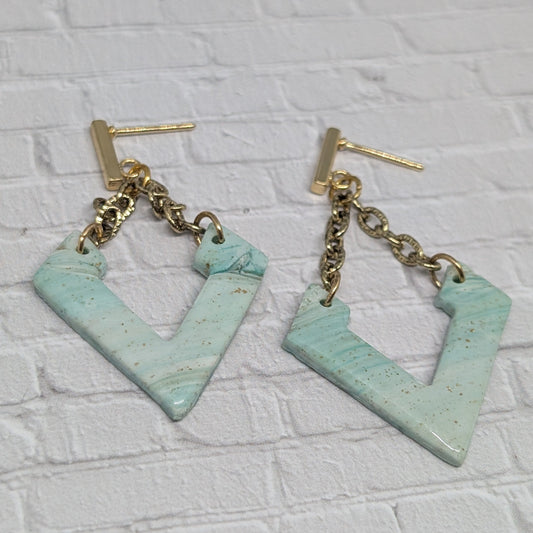 Pair of earrings with gold chains and light blue geometric shapes on a white brick background