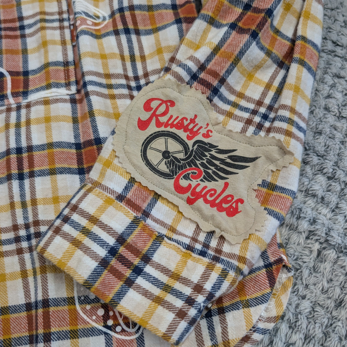 Plaid shirt with 'Rusty's Cycles' logo on a textured surface