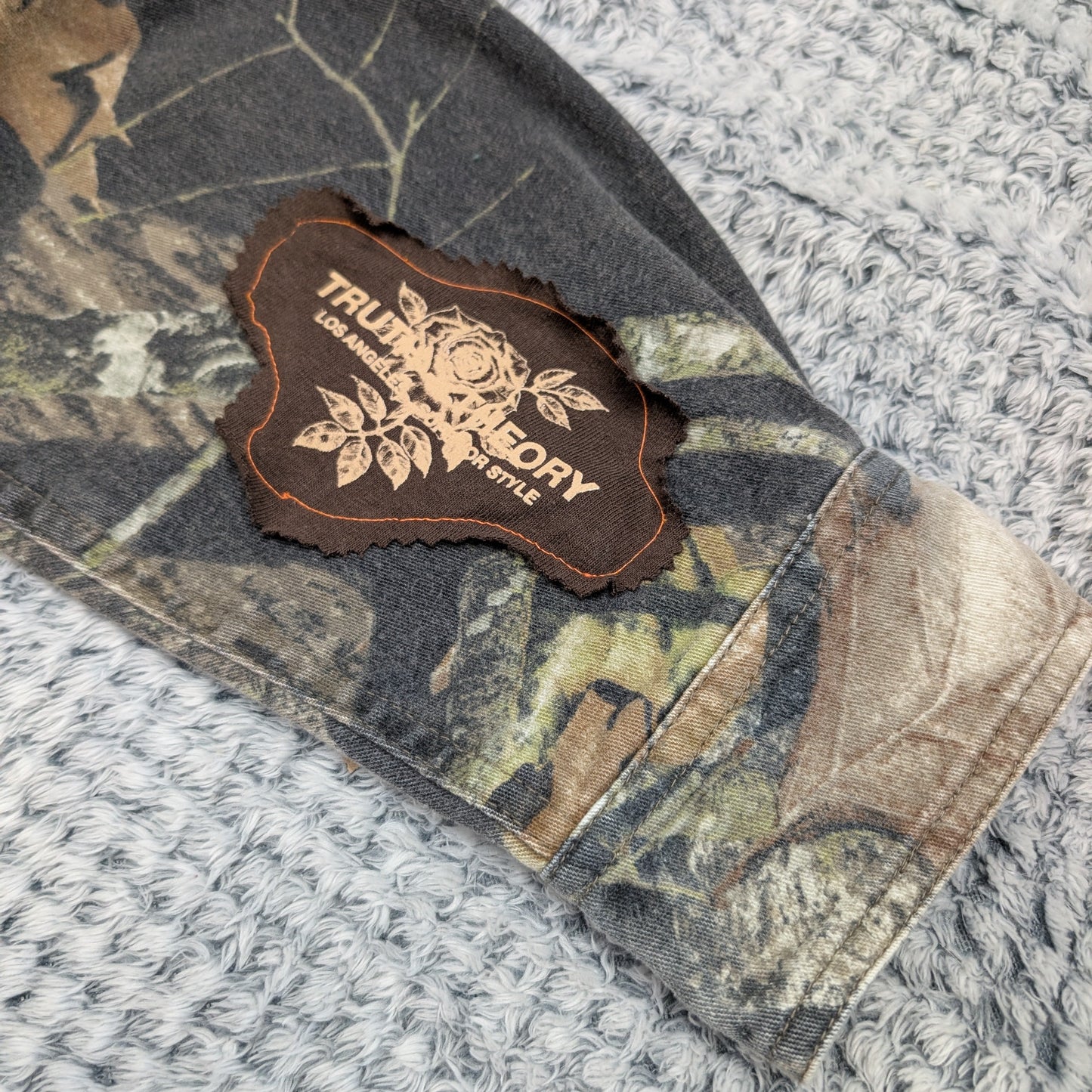 Camouflage-patterned fabric with a leather patch featuring 'True Grit' branding on a textured surface.