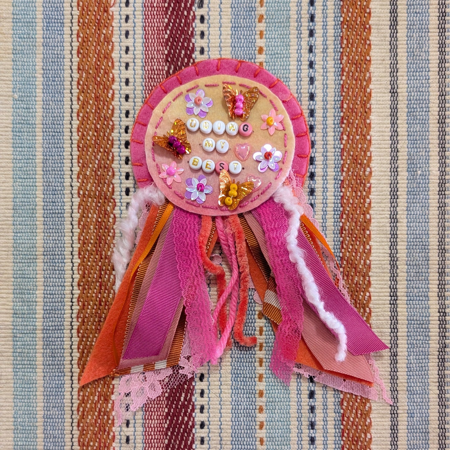 A handmade bohemian-style brooch or pin featuring a circular embellished center with rhinestones and sequins, surrounded by layered pink, orange, and purple satin ribbons, displayed on a striped woven textile background.