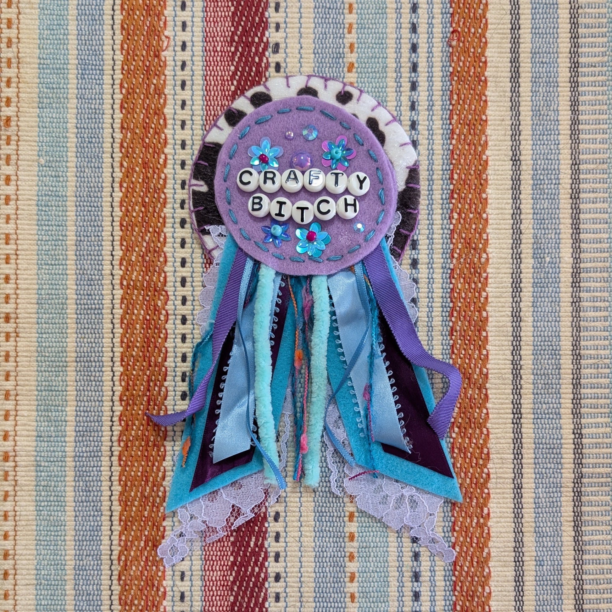 A handmade 'Happy Birthday' ribbon rosette with blue and purple ribbons, displayed on a striped fabric background.