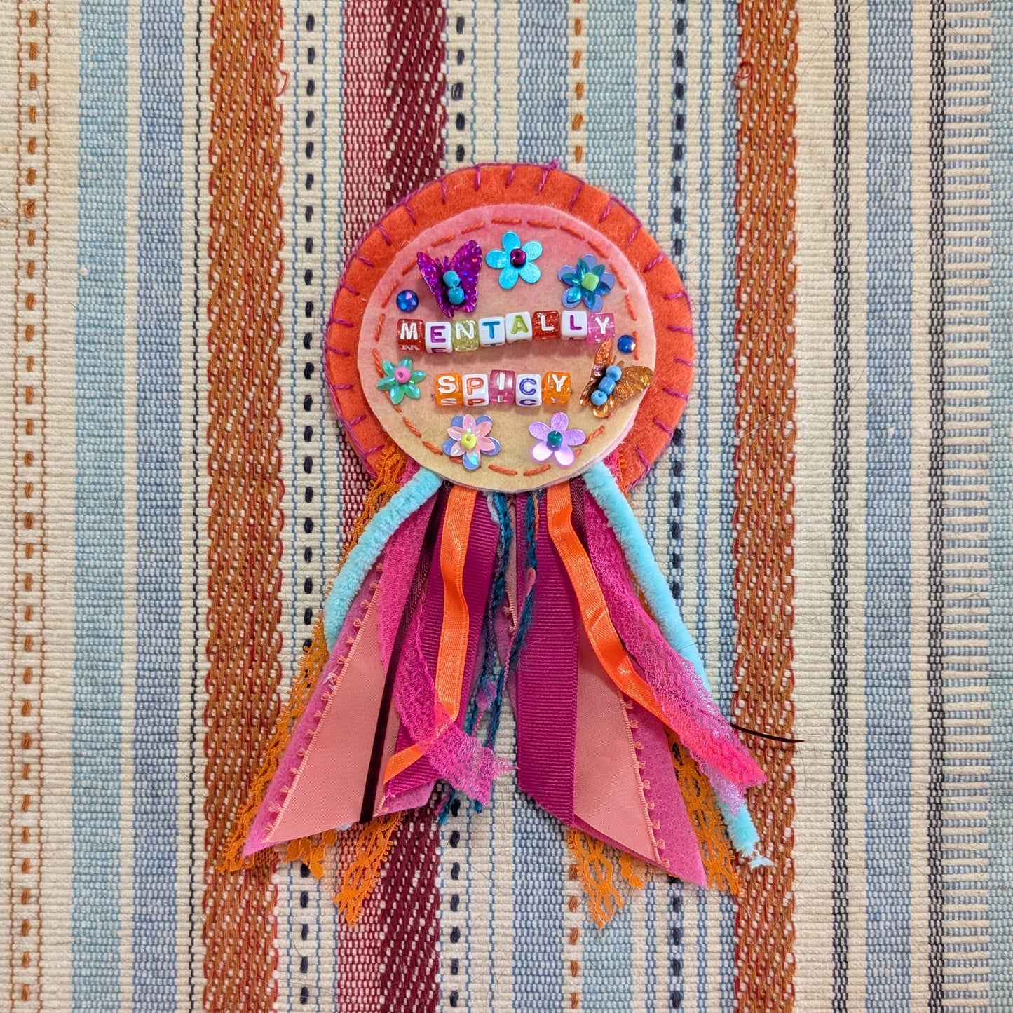 A colorful handmade ribbon award badge with a circular decorated center and multiple pleated paper ribbons, placed on a striped woven textile background.